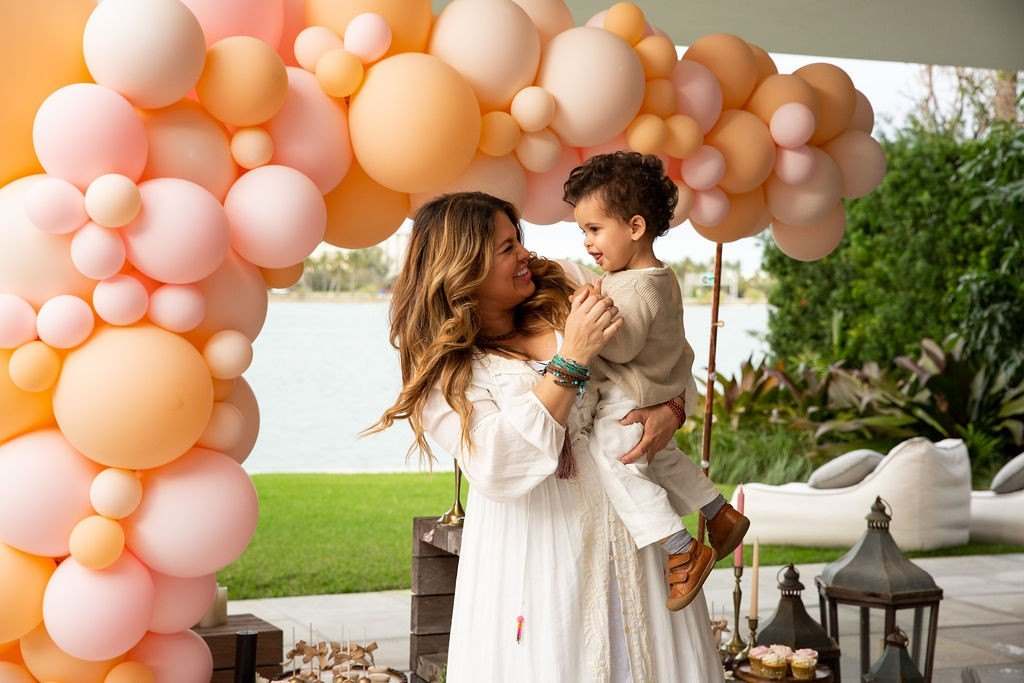 Mother holding her son under a peach balloon arch on a lawn