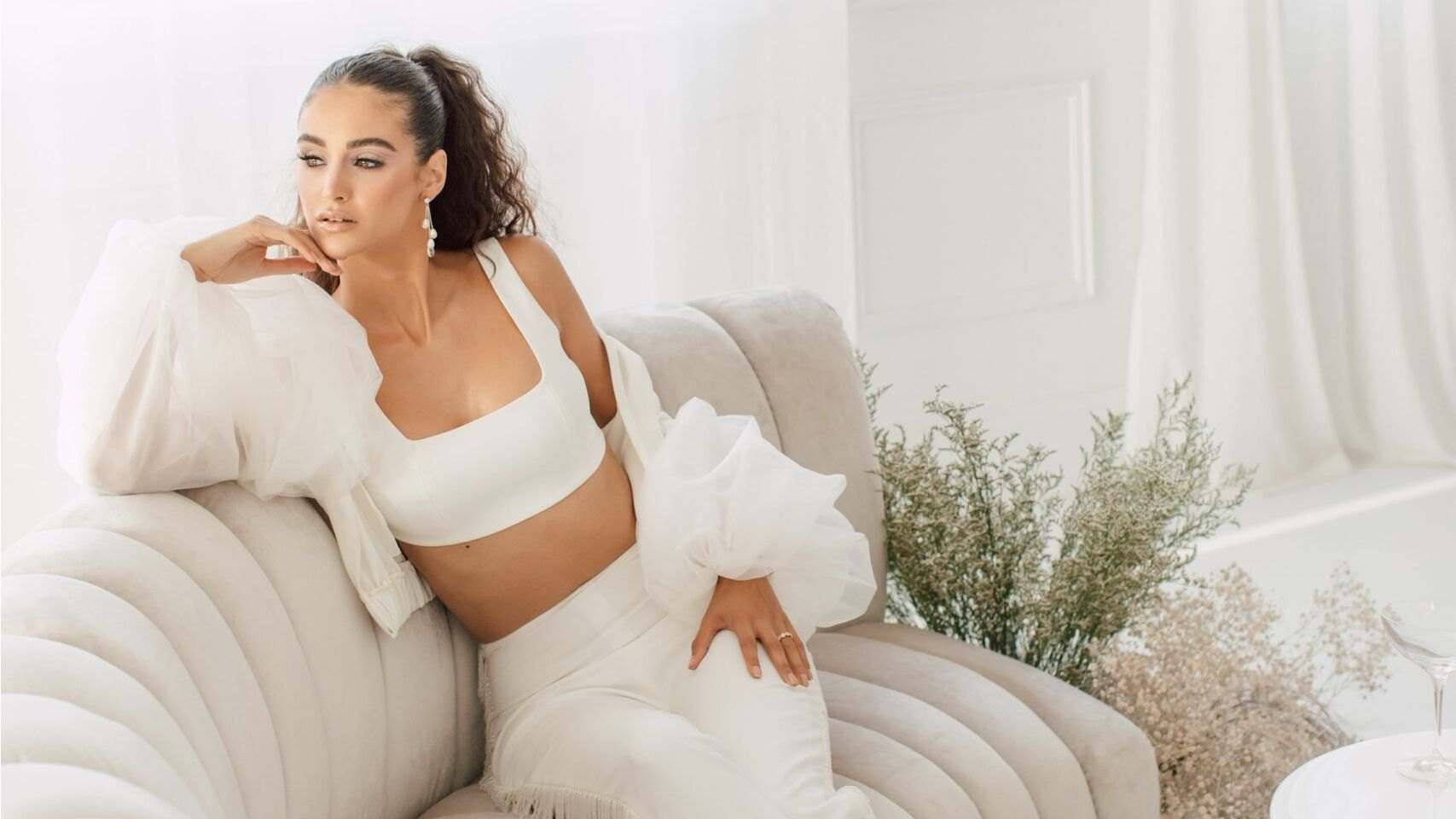 Model in white lounging on a cream sofa beside dried grasses