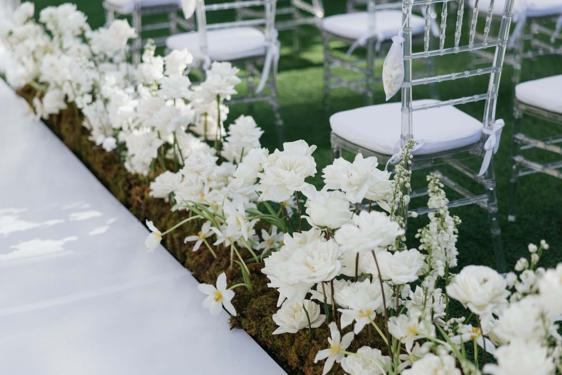 Aisle edged with white orchids and roses on moss beside ghost chairs