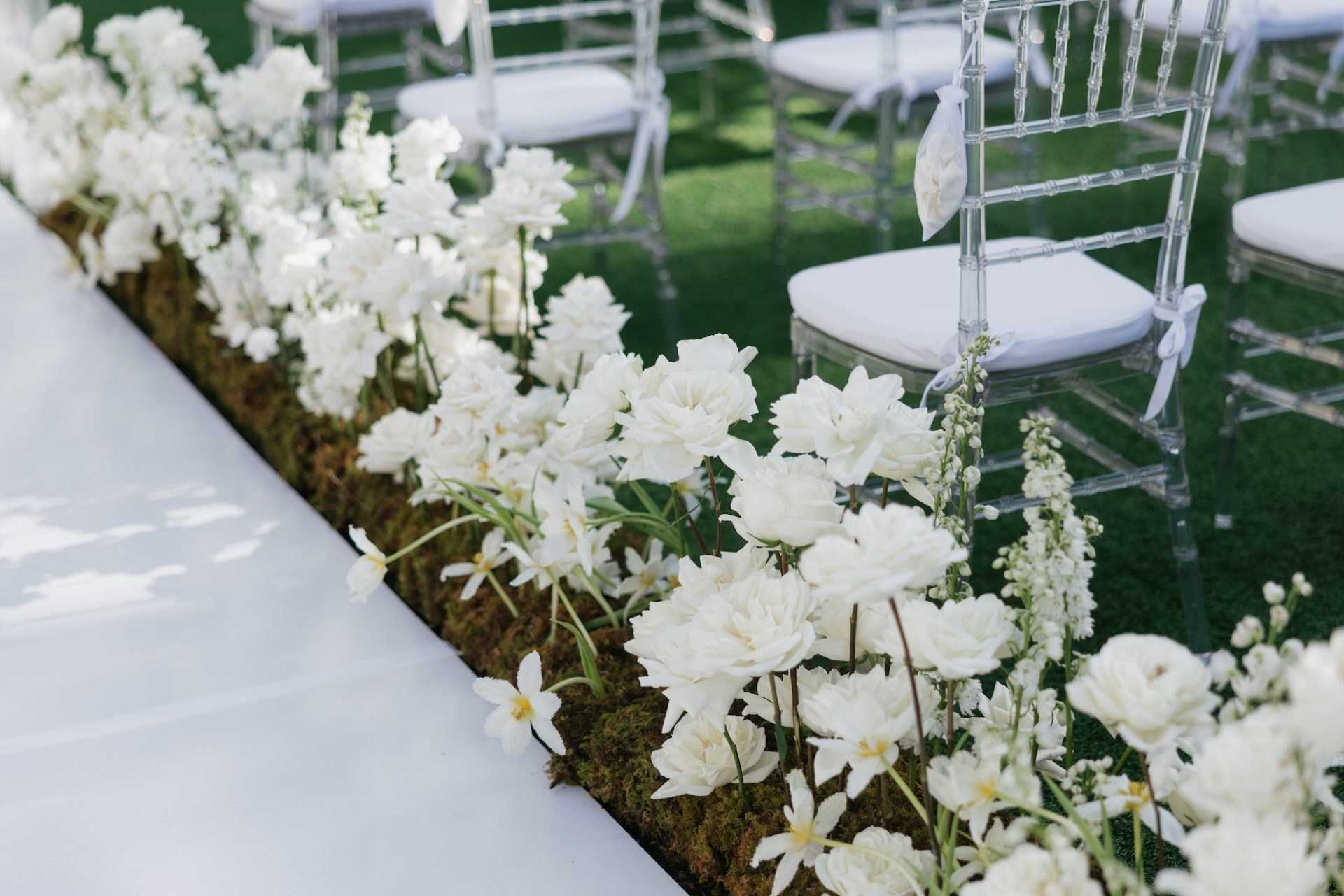 Aisle edged with white orchids and roses on moss beside ghost chairs