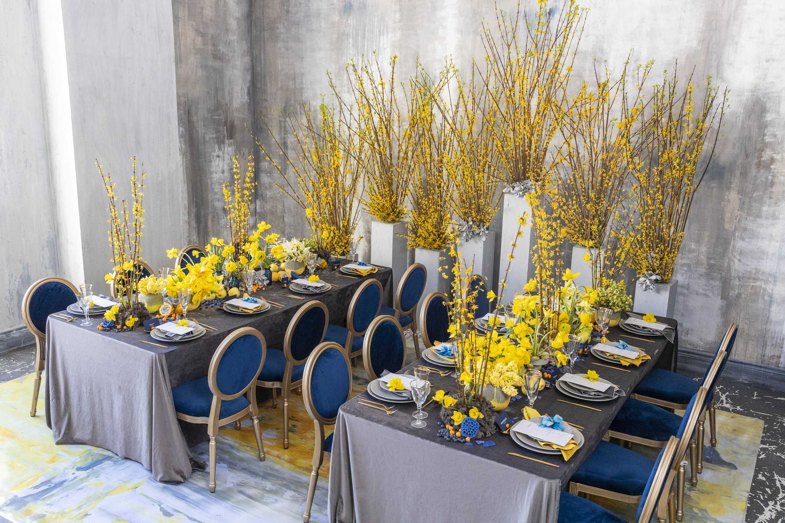 Yellow orchids on tall white vases along a grey banquet with navy chairs